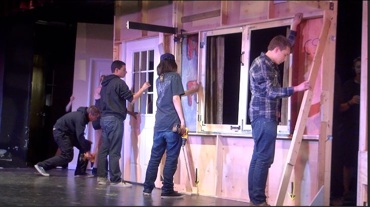 Bühnenarbeiter montieren eine hölzerne Kulisse mit Tür und Fenster für eine Theateraufführung.